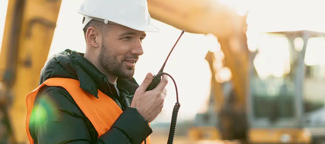 Young engineer talking radio communication (walkie talkie) and wearing a white helmet and construction orange vest. Close up engineers working on a building site with sunny background.